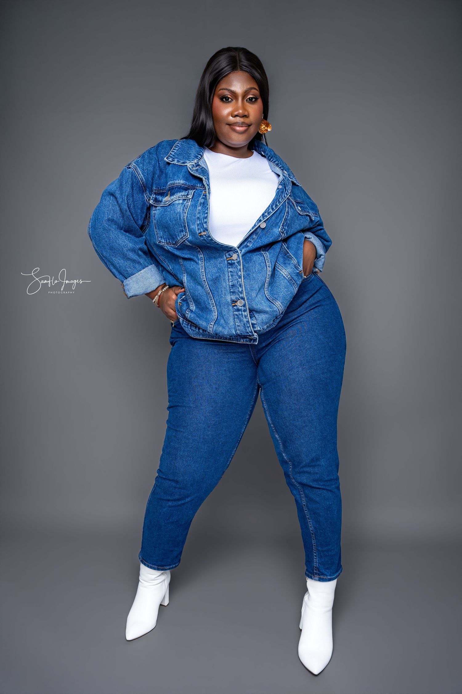 Fashion model wearing double denim outfit with white boots poses confidently against grey background in studio shoot.