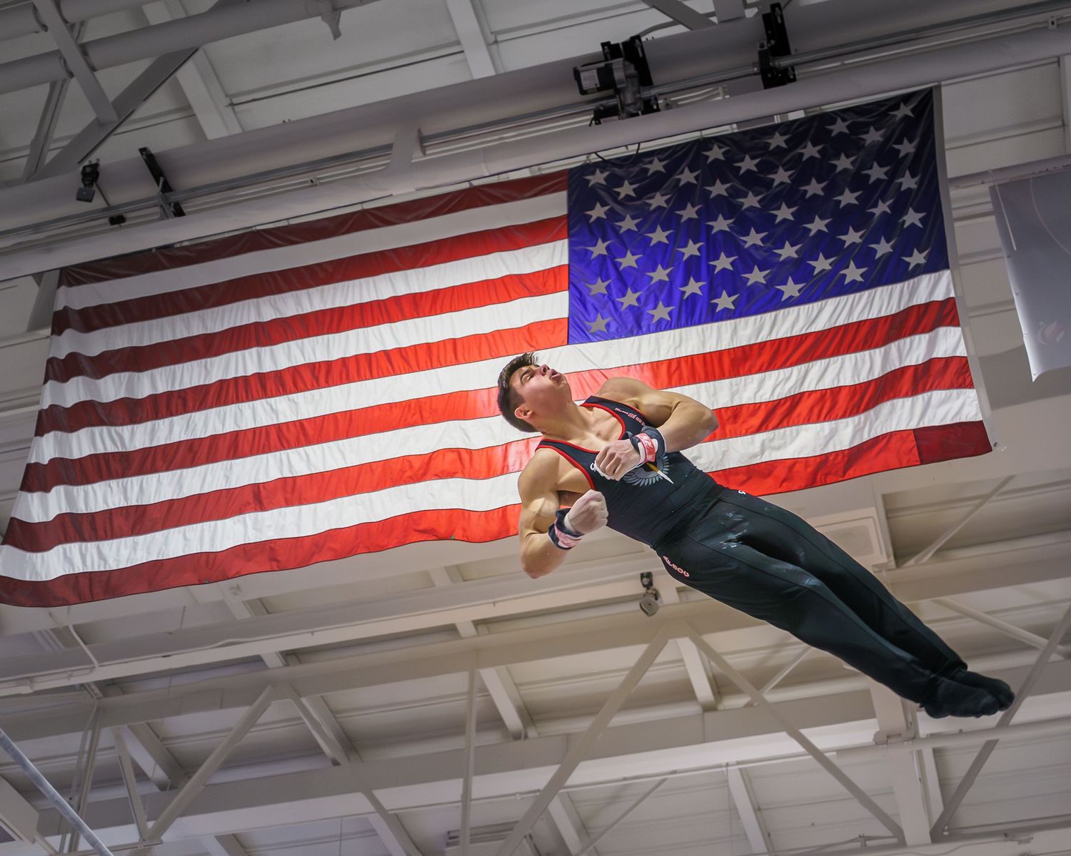 A person performs a mid-air somersault in front of a large American flag hanging on a gymnasium wall.