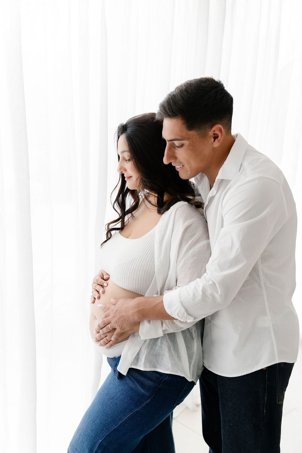 Couple in white and denim embracing during maternity photoshoot against bright window backdrop.