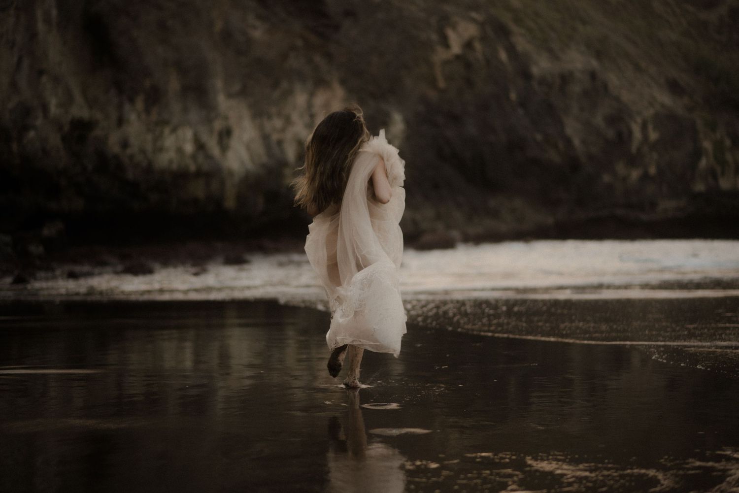 A person in a flowing white dress stands alone on a misty beach at dusk creating a dreamy ethereal scene.