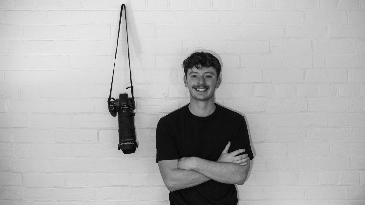 Black and white portrait of photographer Jayden Threadgold standing against a brick wall with a camera hanging nearby for Threadgold Media