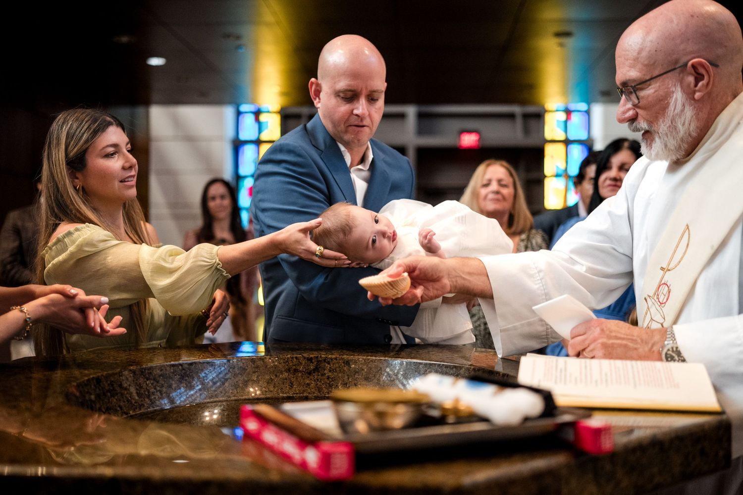 Priest performing Catholic baptism ceremony with parents holding infant over baptismal font in church.