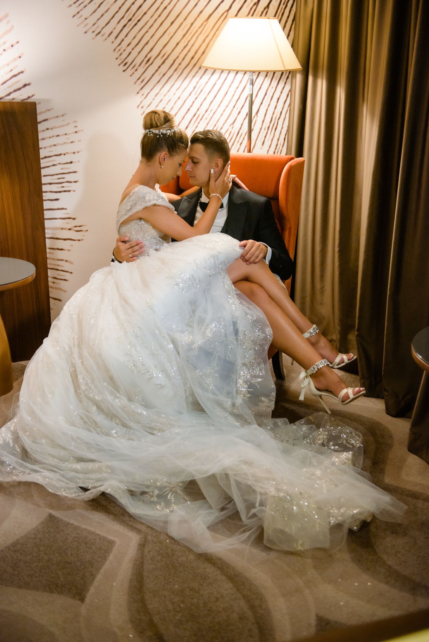 Tender moment between newlyweds sitting in orange chair with elegant white wedding dress.