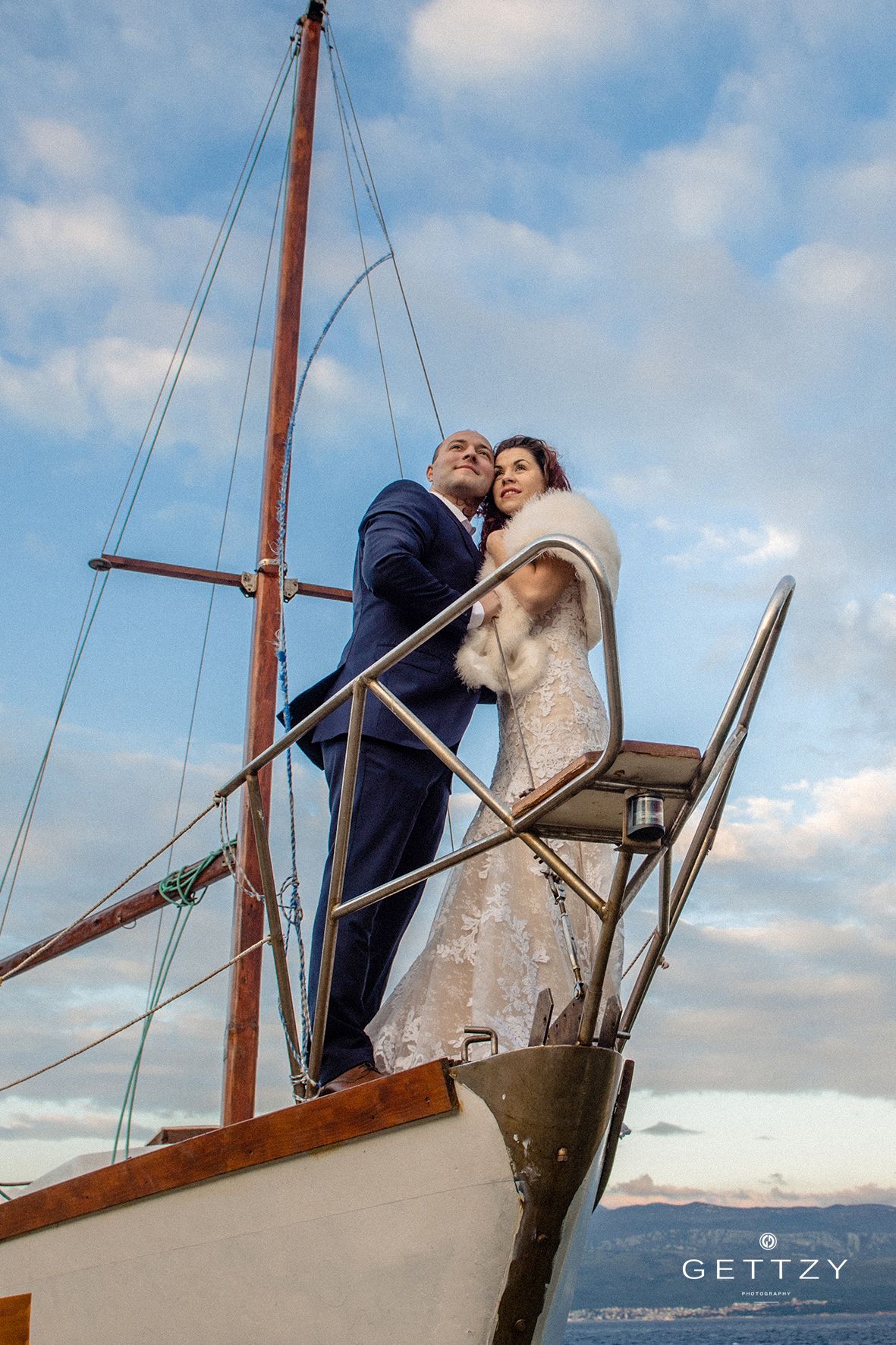 Sailing vessel wedding photo with couple embracing on bow against cloudy sky.