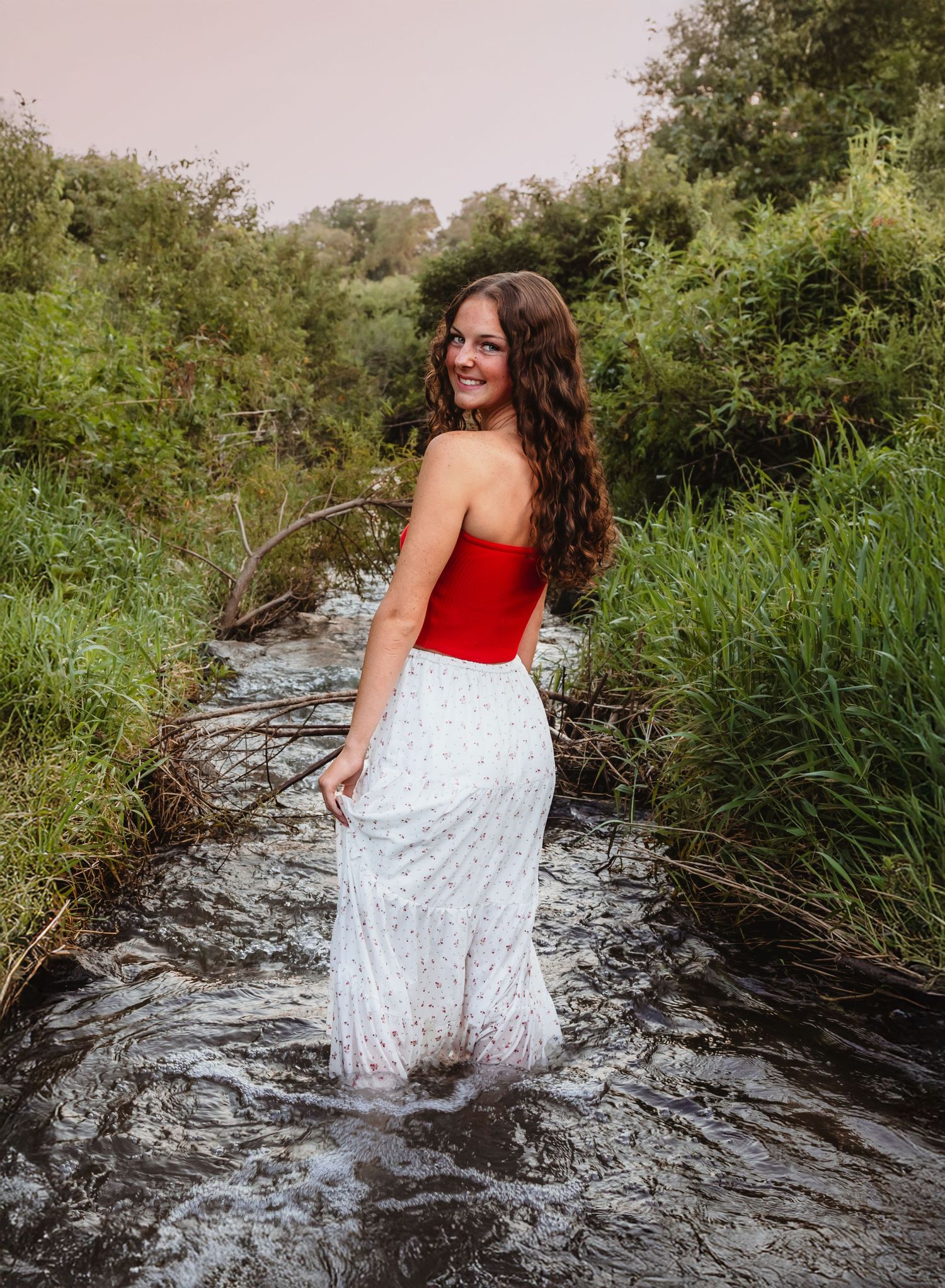 A senior girl in a red and white dress splashes playfully in a flowing creek surrounded by lush greenery.