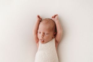 A newborn baby in white stretches with arms raised against a light background.