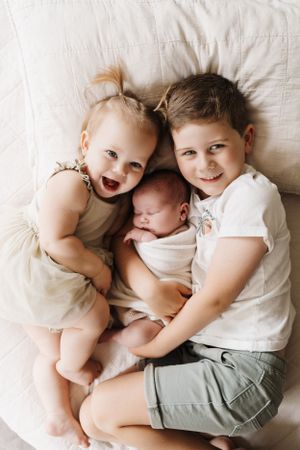 Three young siblings lie together on white bedding with joyful expressions.