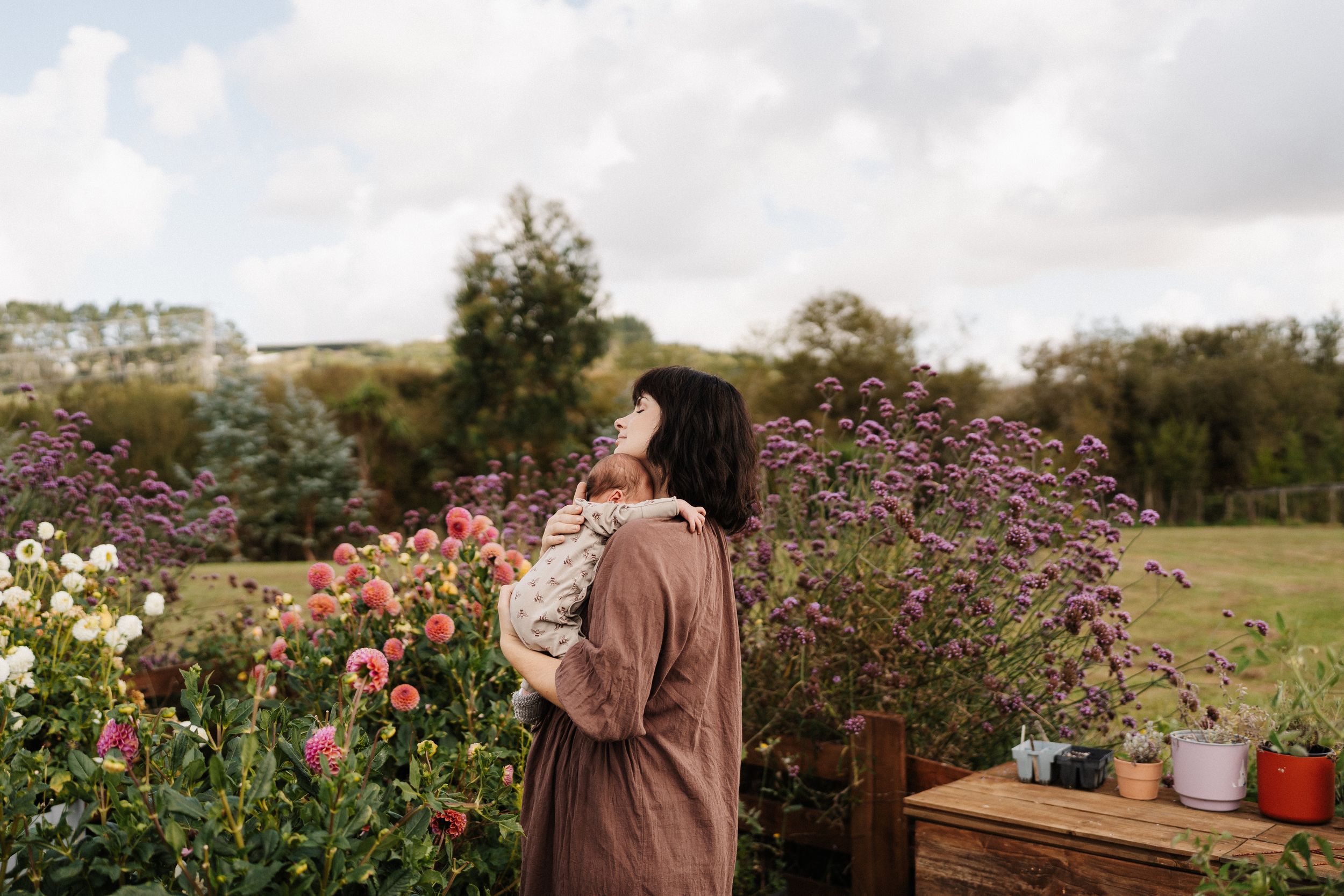 A person in a brown jacket stands in a lush garden filled with blooming flowers and greenery on a sunny day.