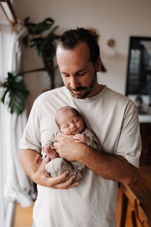 A tender moment shows someone in a white t-shirt holding and cradling a sleeping newborn baby in a home setting.