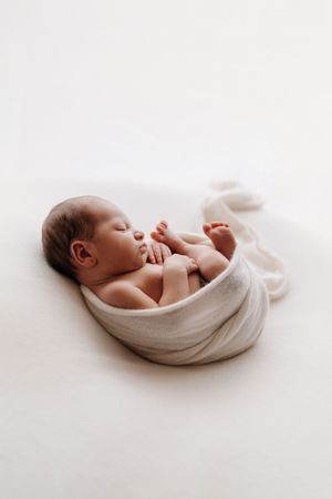 Peaceful sleeping newborn wrapped in soft white fabric against light background during professional photoshoot.