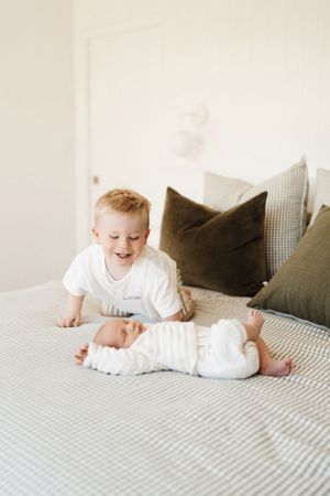 Baby in white clothing playing and smiling on a striped bed with pillows.