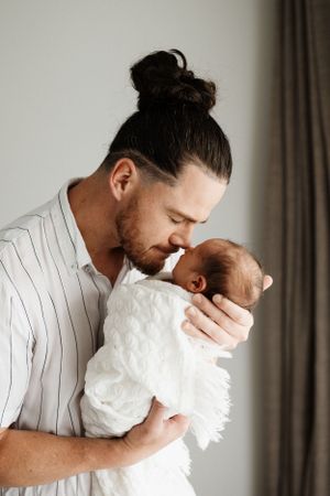 Father in striped shirt tenderly holds sleeping newborn wrapped in white.