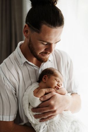 Father in white striped shirt cradles sleeping newborn against window light.