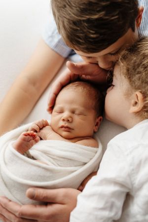 Siblings share intimate moment with sleeping newborn wrapped in white.