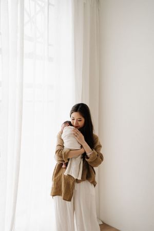Woman in tan cardigan embraces white pillow near bright window.