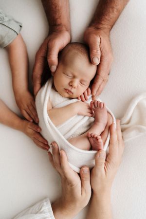 Overhead view of gentle hands cradling sleeping newborn in white wrap.
