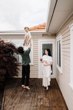 Family playing together on a wooden deck outside their beige house with decorative burgundy shrub.