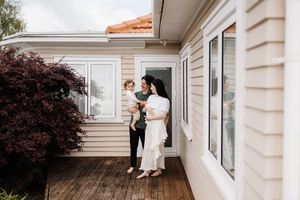 A couple sits together on a white porch railing outside a house with cream siding and dark landscaping.