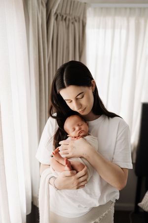 A mother in a white t-shirt tenderly holds and gazes down at her sleeping newborn baby near a window with light curtains.