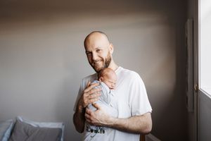 A person in a white shirt holds a newborn baby while standing near a window in a softly lit room.