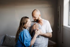 Couple in casual clothing shares tender moment while holding newborn baby in sunlit room.