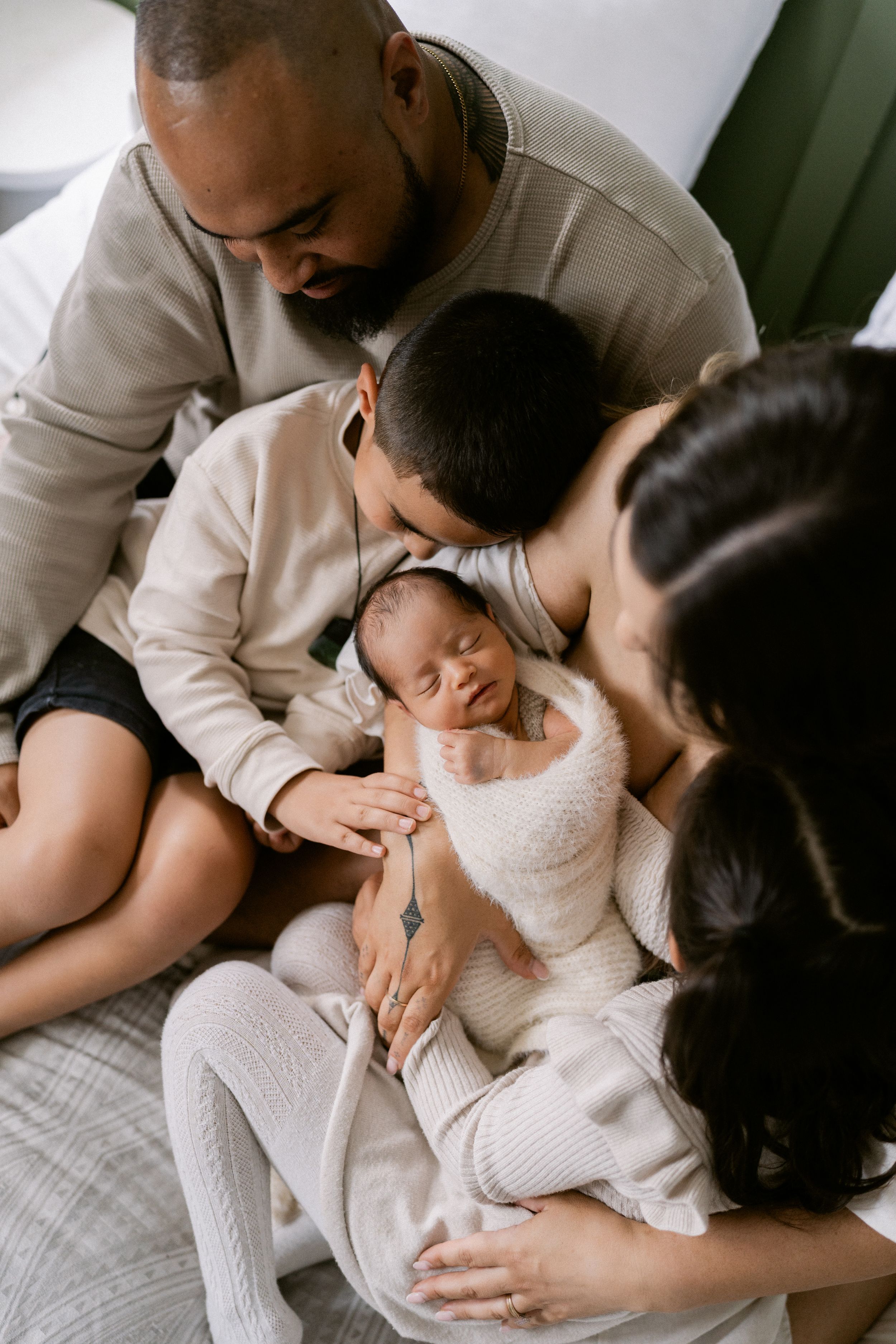 A loving family of four gathers closely, admiring their sleeping newborn baby wrapped in a white blanket.
