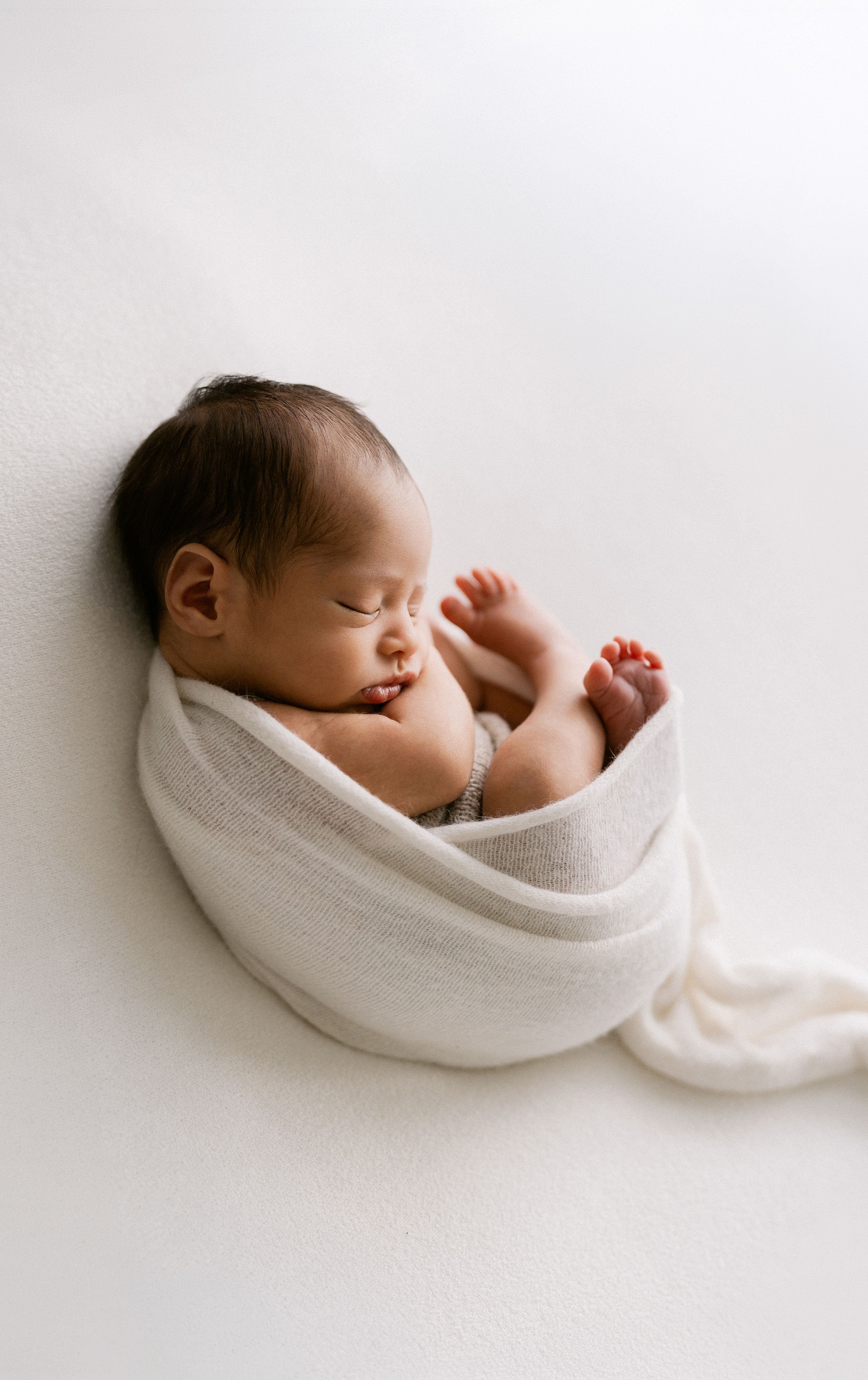 Sleeping newborn baby swaddled in a soft white wrap, peacefully resting against a white background.