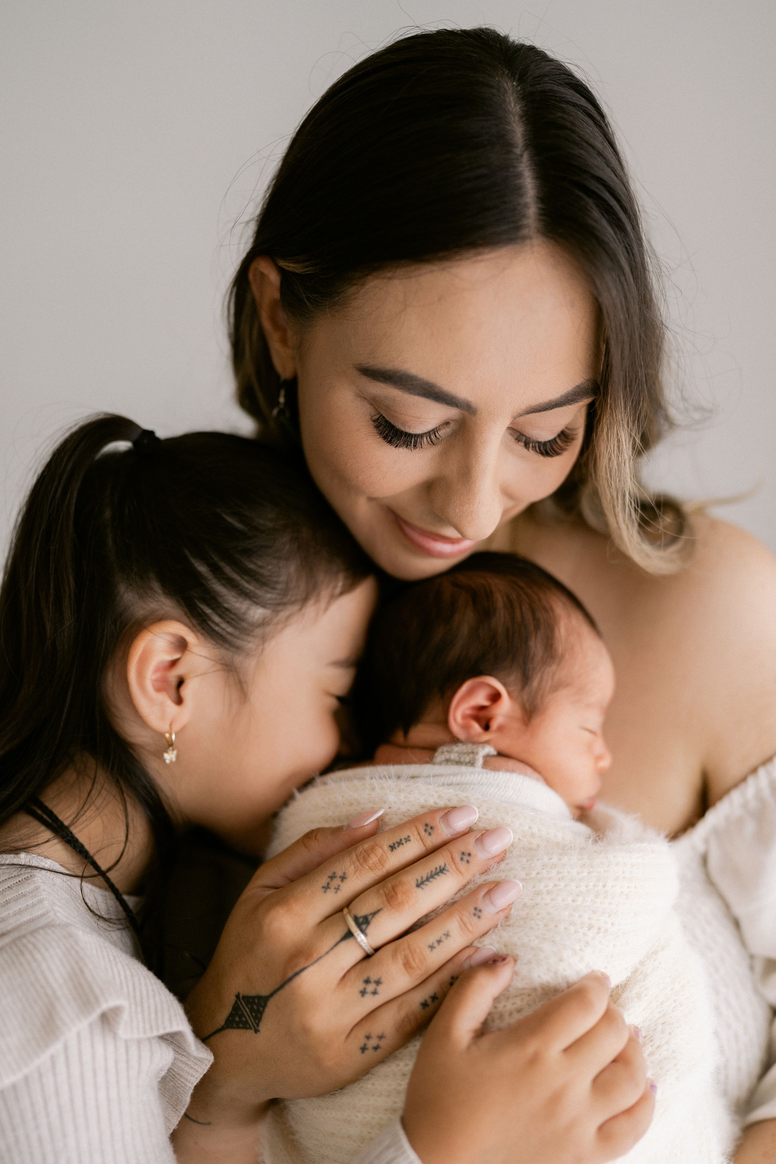 A mother and young daughter lovingly cradle a swaddled newborn baby in a soft, bright studio setting.