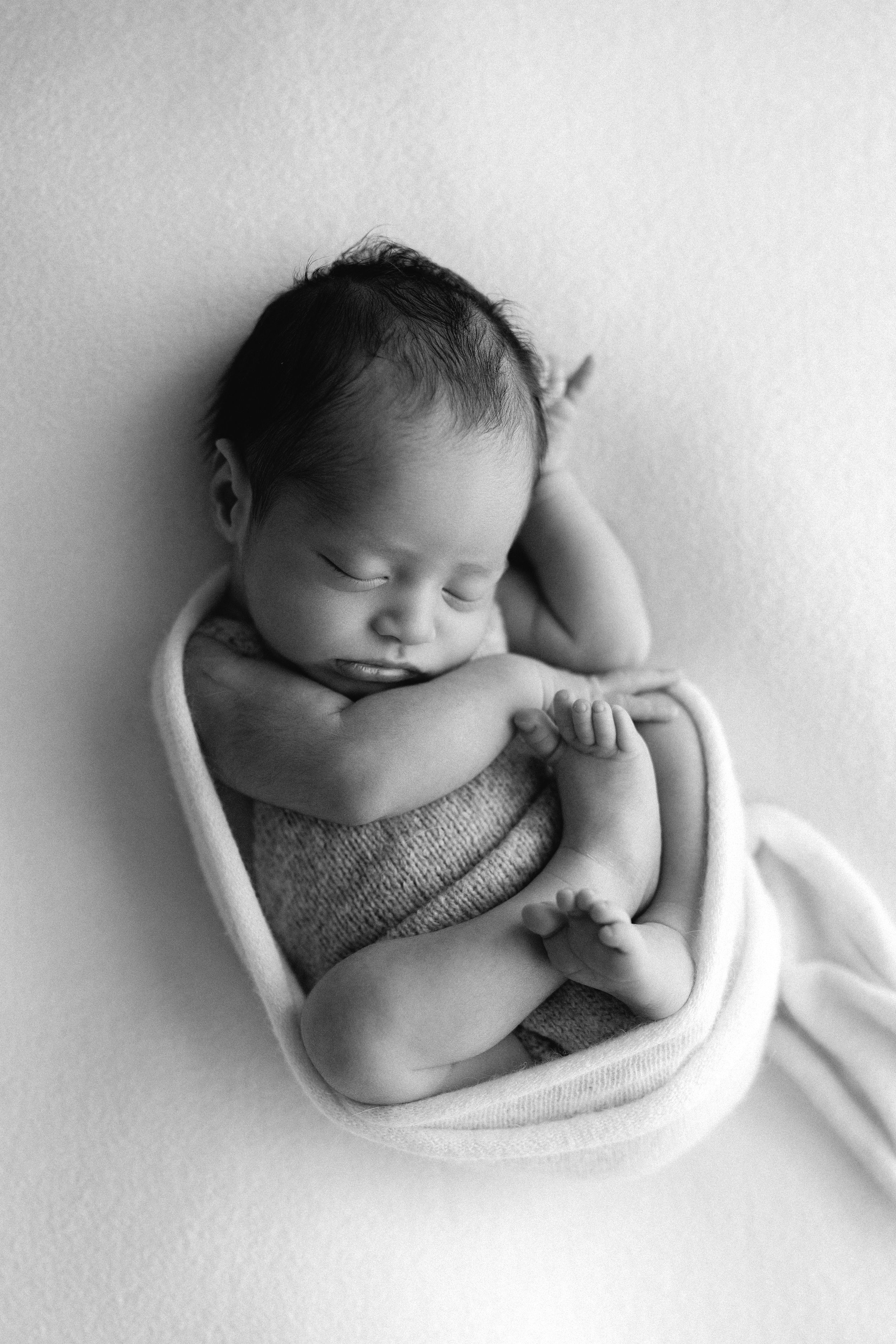 Black and white photo of a sleeping newborn baby curled up snugly in a small bowl wrap.
