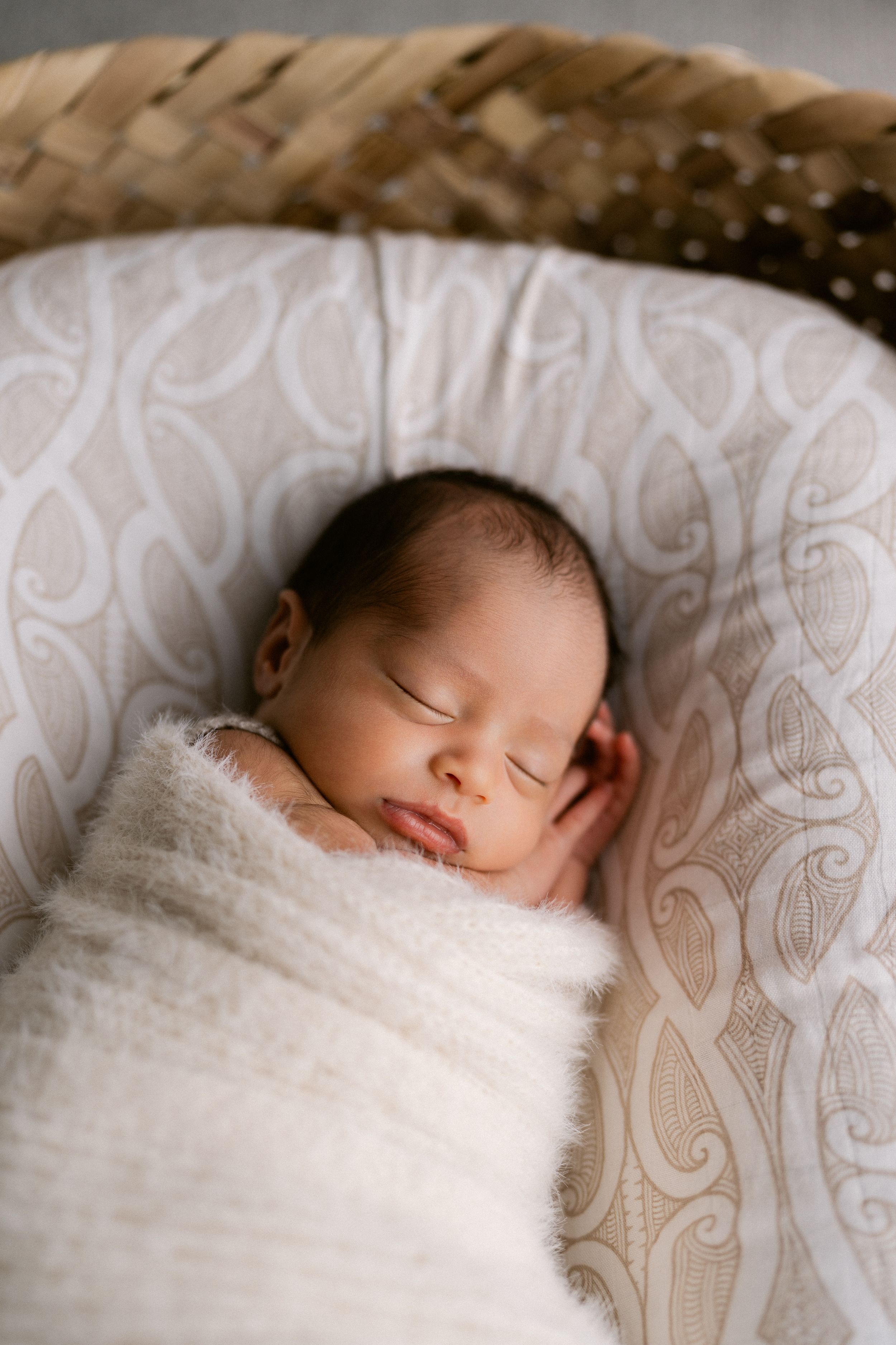 Peaceful newborn baby swaddled in white wrap sleeping in a woven bassinet with neutral patterned lining.