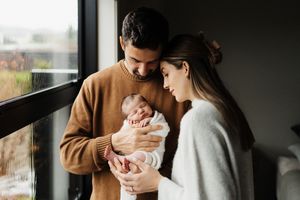 Parents lovingly hold and care for their newborn baby in a series of intimate indoor portraits by a window.