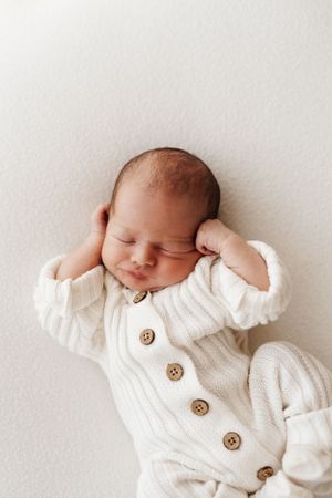 Sleeping newborn stretches in white knit cardigan with wooden buttons.