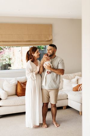 Couple in white and beige outfits holding their newborn baby in bright living room.