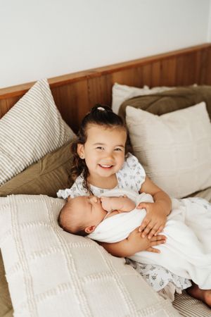 Two siblings share a tender moment on white bedding as older sister cuddles with sleeping baby.