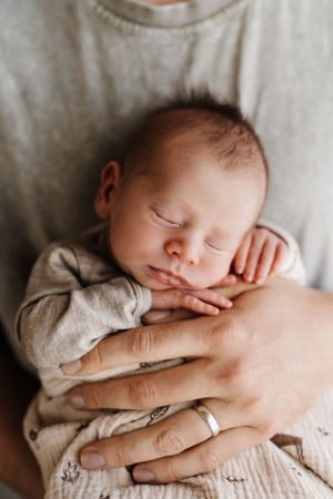 A peaceful sleeping newborn wearing a light gray onesie is cradled in gentle hands with a wedding ring visible.