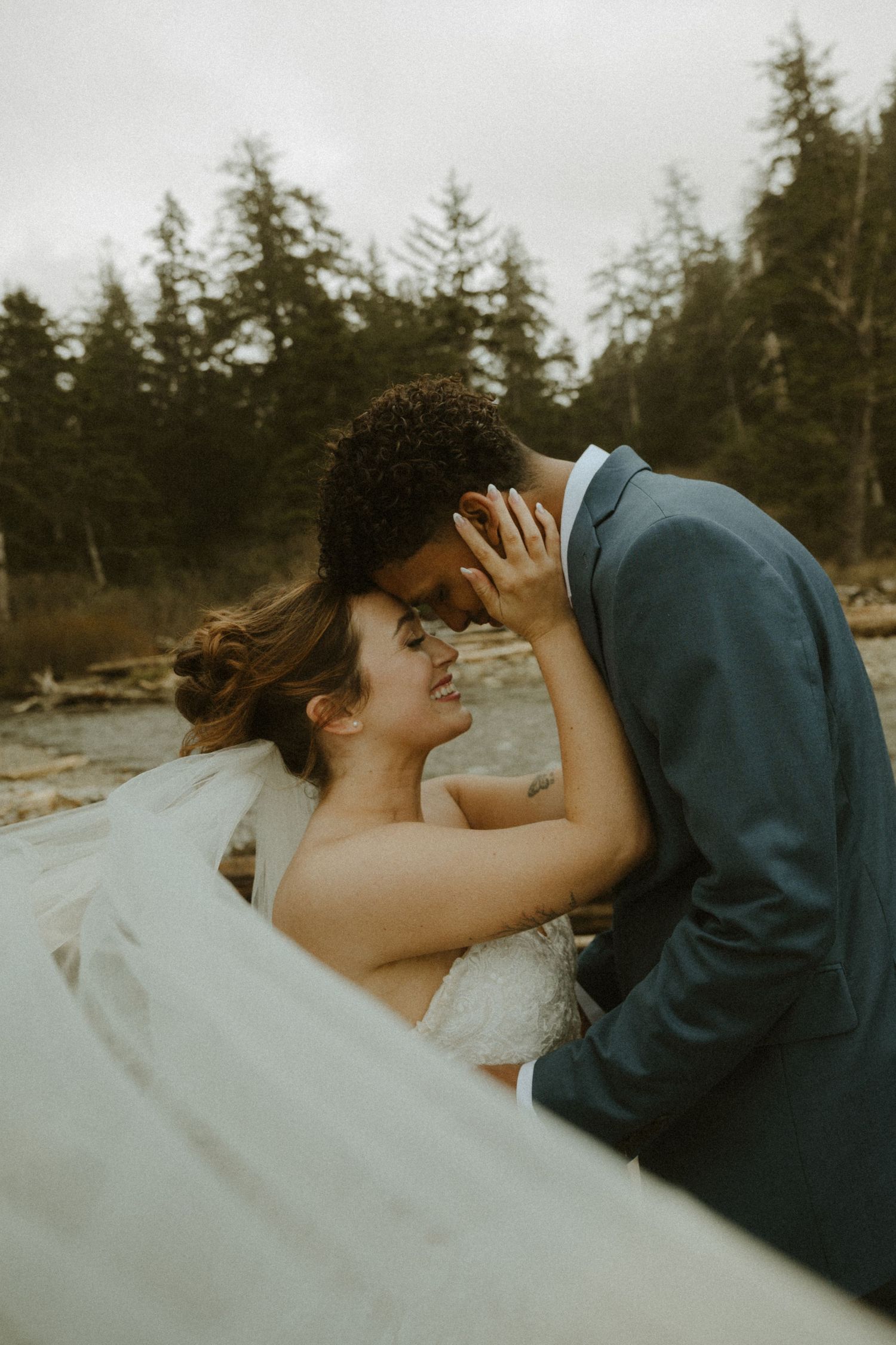 A couple shares a romantic kiss on a beach with evergreen trees in the background.