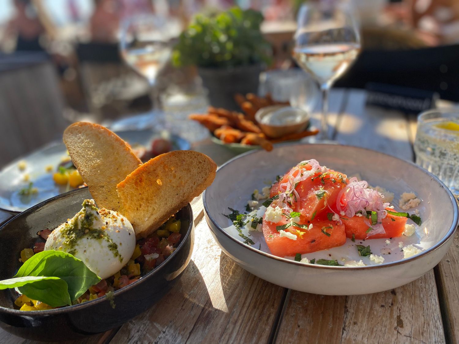 Two gourmet dishes with burrata and salmon served outdoors on wooden table with wine glasses.