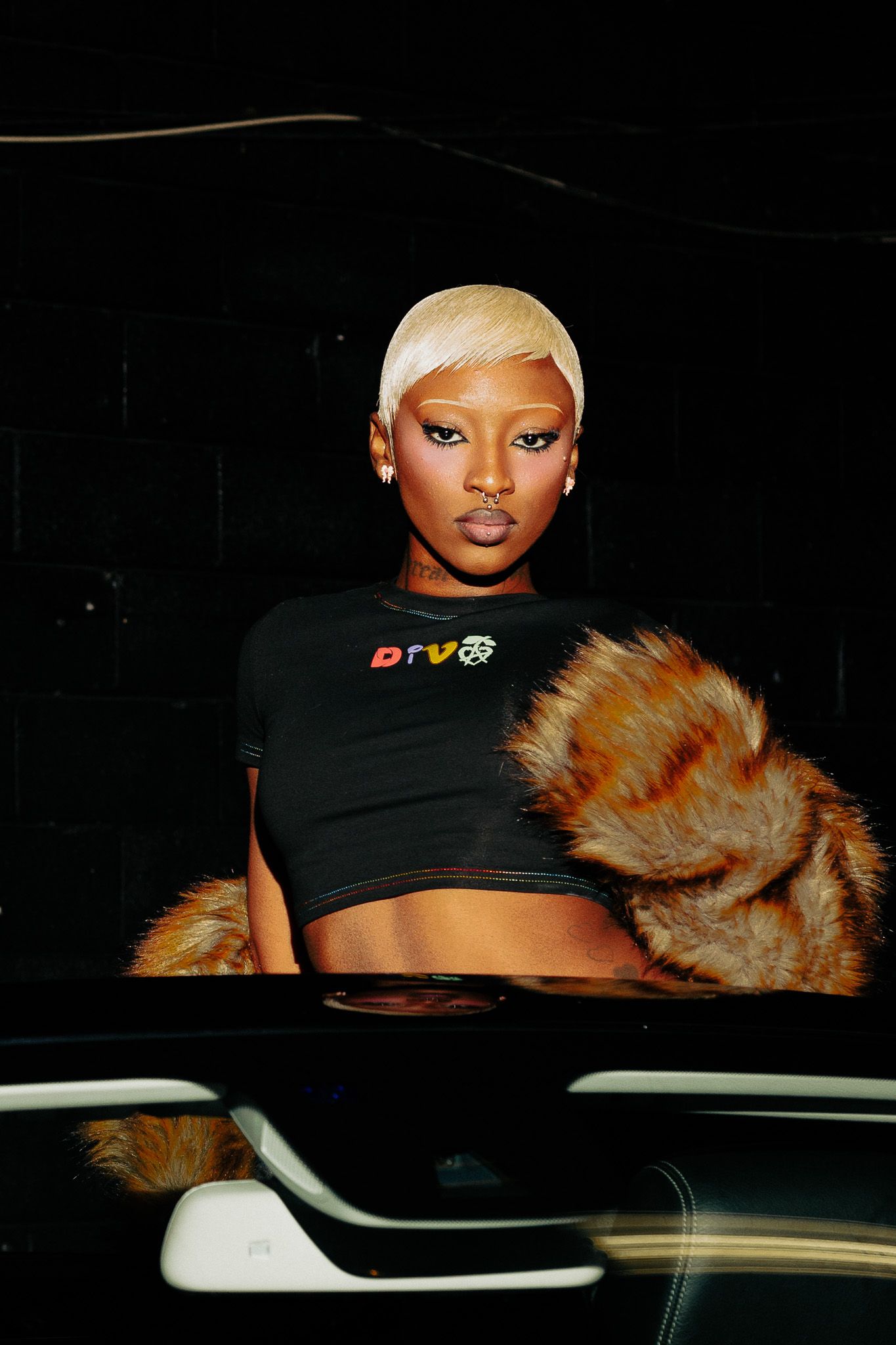 A person wearing a black cap and fur accessory poses in a dark car interior.
