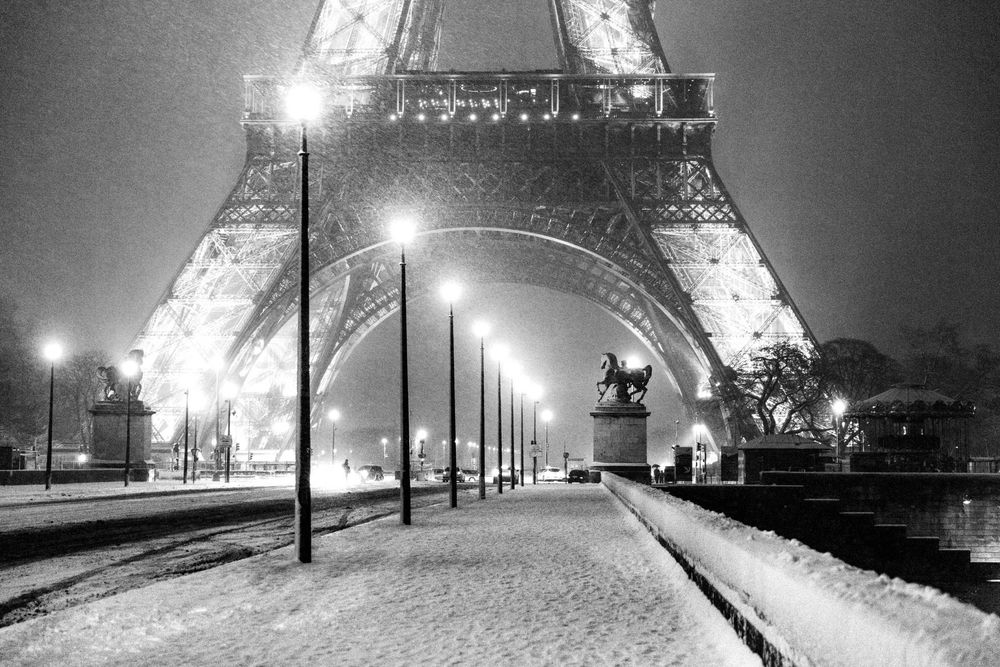 Black and white photo of the Eiffel Tower at night in winter, with snow-covered paths and glowing street lamps.