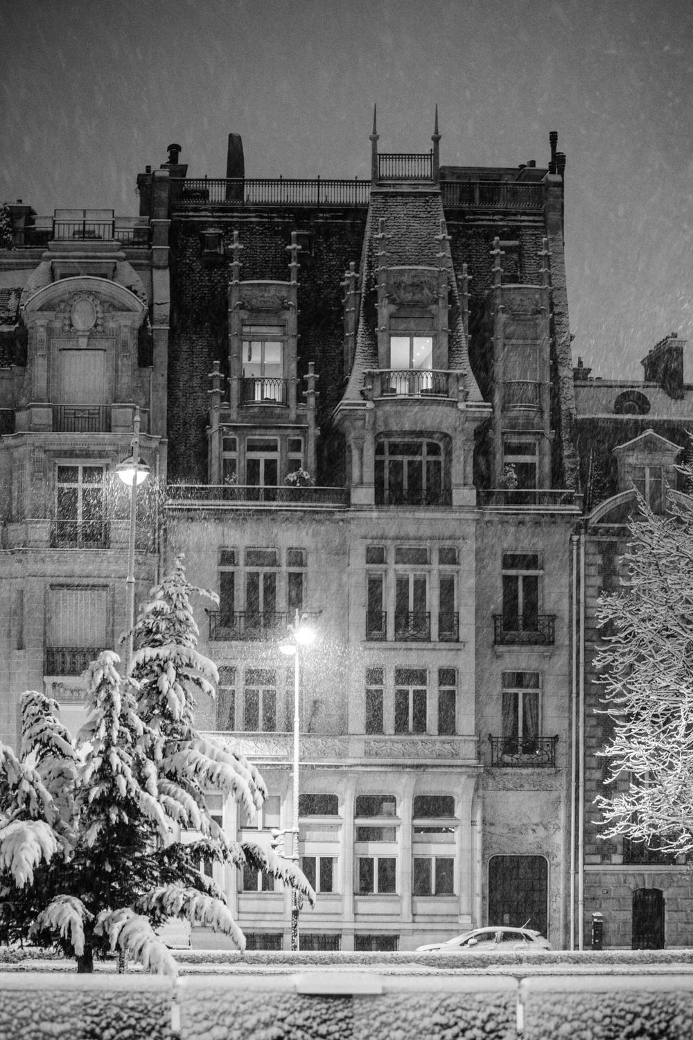Black and white photo of a grand Parisian building covered in snow at night, illuminated by street lamps.