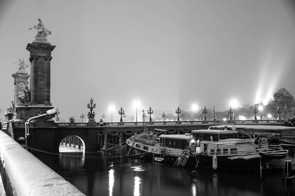 Black and white view of Pont Alexandre III bridge with moored boats and glowing streetlights in snowy Paris night.