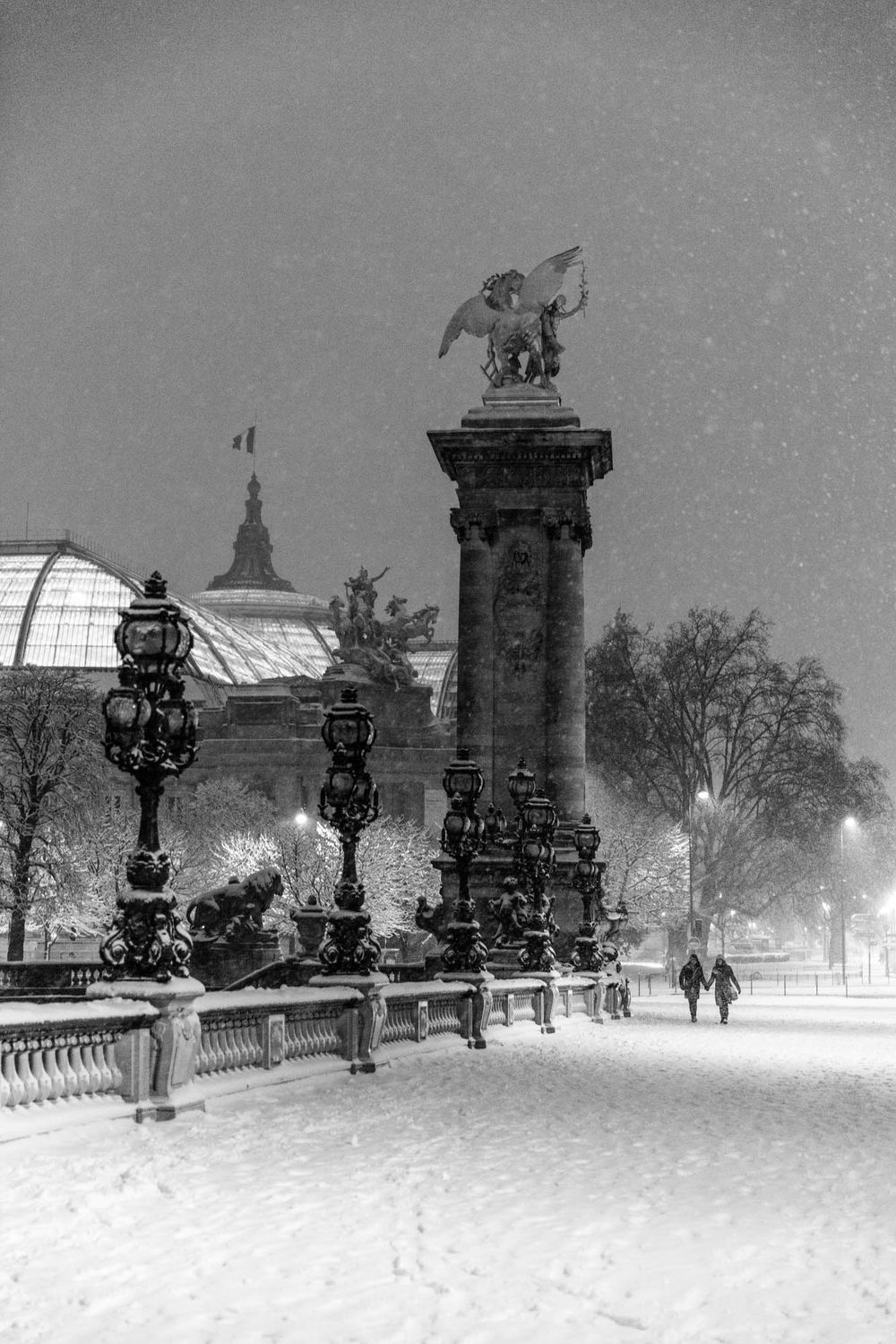 Black and white photo of Pont Alexandre III bridge in Paris during heavy snowfall at night with Grand Palais in background.