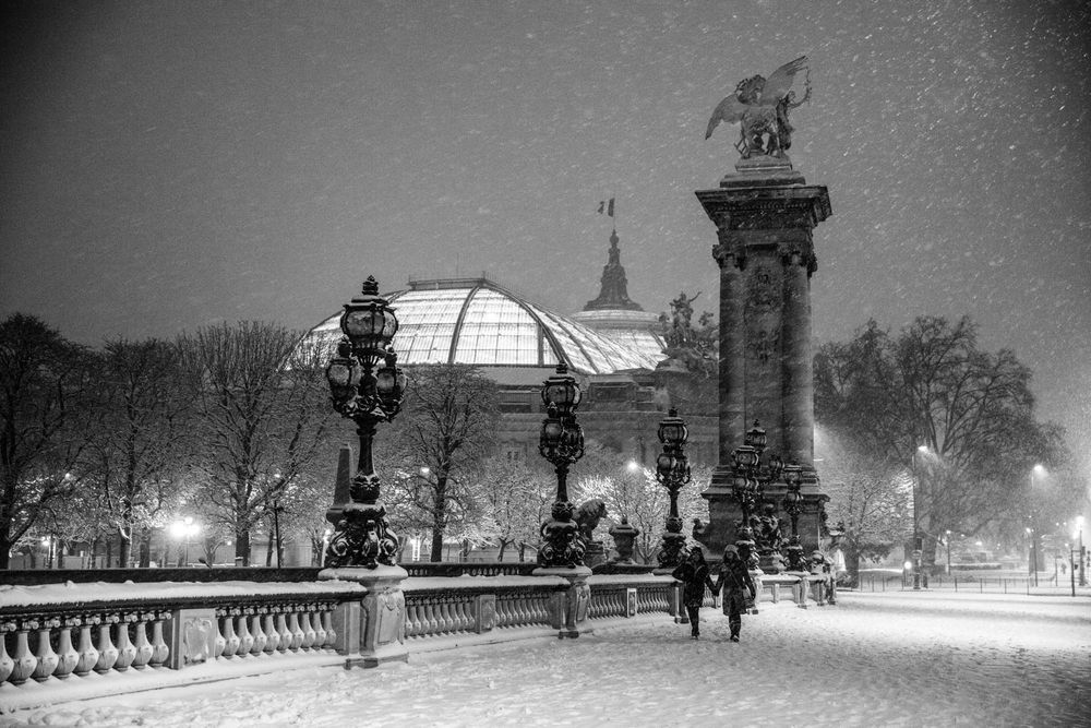 Black and white photo of snowy Paris night near Grand Palais with glowing street lamps and pedestrians walking.