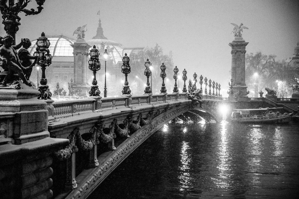 Black and white photo of the ornate Pont Alexandre III bridge in Paris at night, reflecting lights on the Seine River.