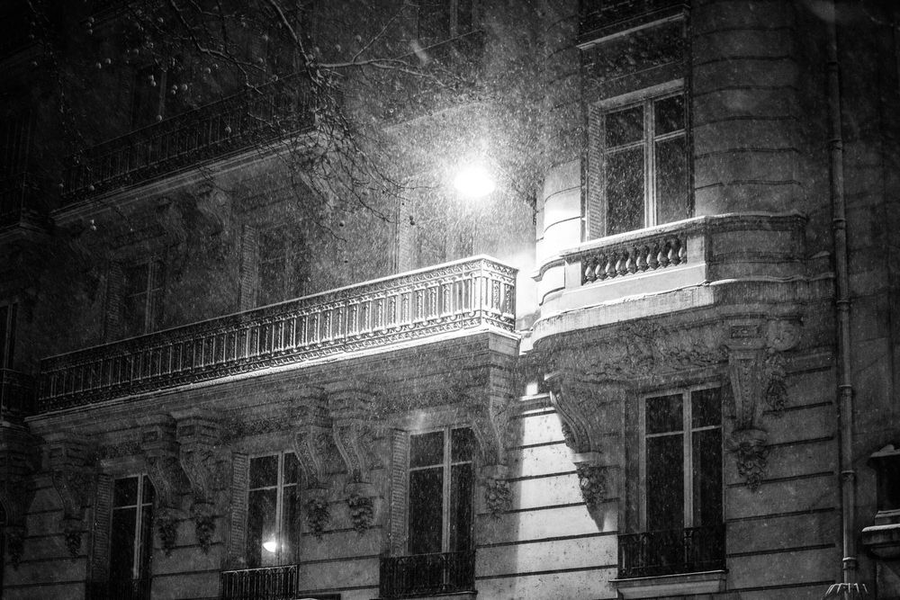 Black and white night photo of a classic Parisian building facade with a glowing street lamp during snowfall.