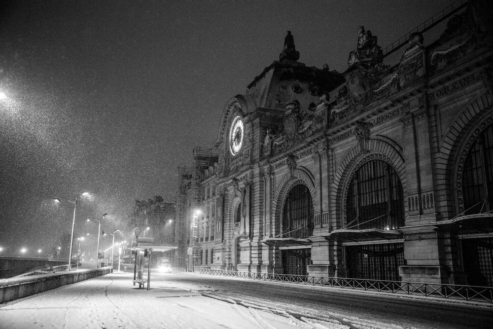 Black and white photo of a grand historic train station at night during a snowstorm with glowing street lamps.