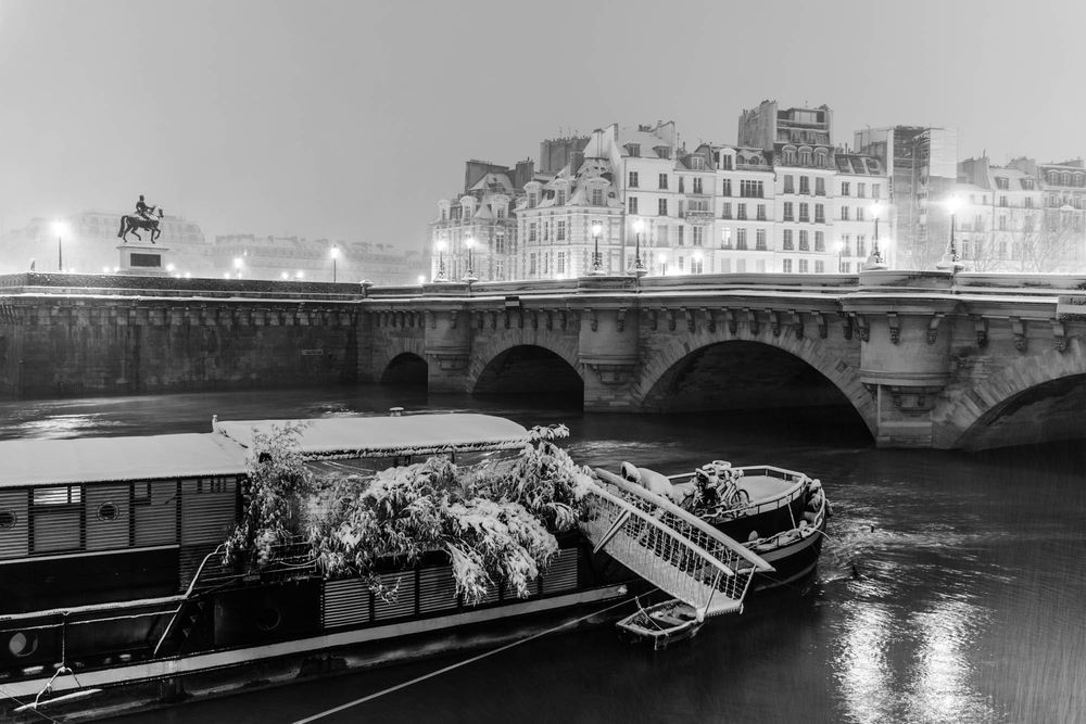 Black and white night photo of a barge on the Seine River with Pont Neuf bridge and Paris buildings behind.
