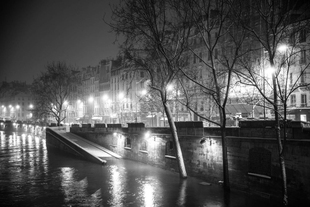 Black and white night photo of a rainy Parisian riverside with glowing street lamps reflecting on wet cobblestones.