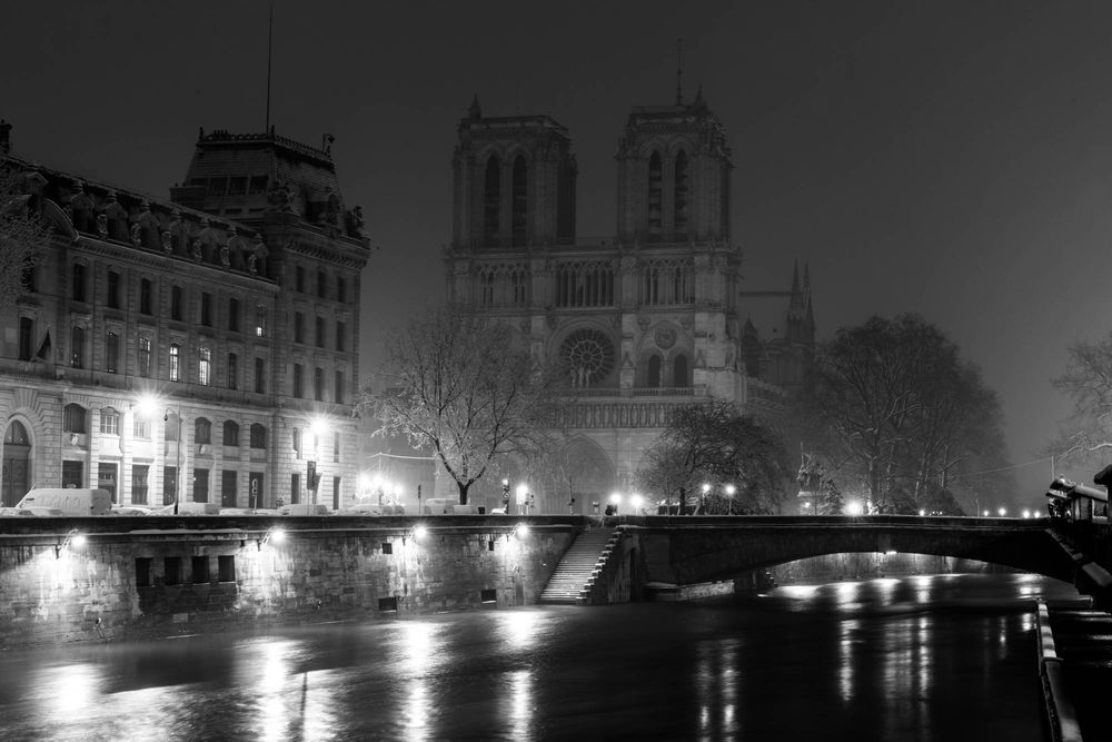 Black and white night photo of Notre-Dame Cathedral in Paris reflected in the Seine River with glowing street lights.