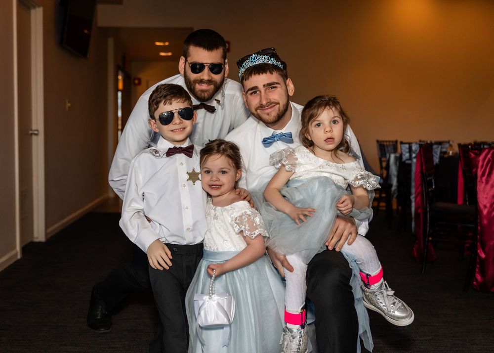 Two men in bow ties pose with three young children dressed formally at a wedding celebration.